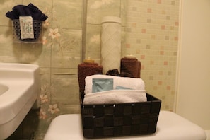 A bathroom with a toilet, a basket containing toiletries, and a towel rack with towels. - Wakaw Lodge Motel (Wakaw)