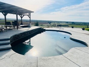 A swimming pool with a stone edge, surrounded by a patio area with seating and a covered section. - Hillside Acres Tiny Home (Minkler)