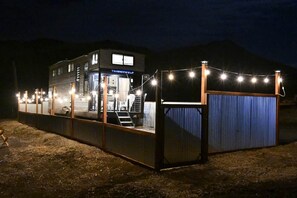 A building with a visible entrance, a staircase leading to the upper floor, and a fenced area with string lights. - Hillside Acres Tiny Home (Minkler)