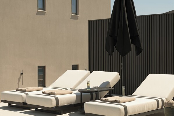 A rooftop lounge area with three sunbeds, a table with a bottle and glass, and a black umbrella. - Neme Slow Living Retreat (Rethymno)