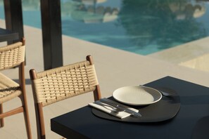A dining table with a plate, cutlery, and a napkin, set against a backdrop of a swimming pool and a chair with a woven backrest.