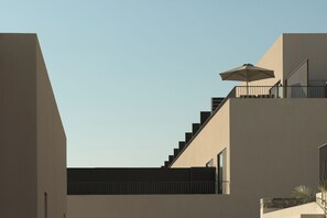 A modern building with a staircase leading to a balcony, a large umbrella, and a clear sky. - Neme Slow Living Retreat (Rethymno)