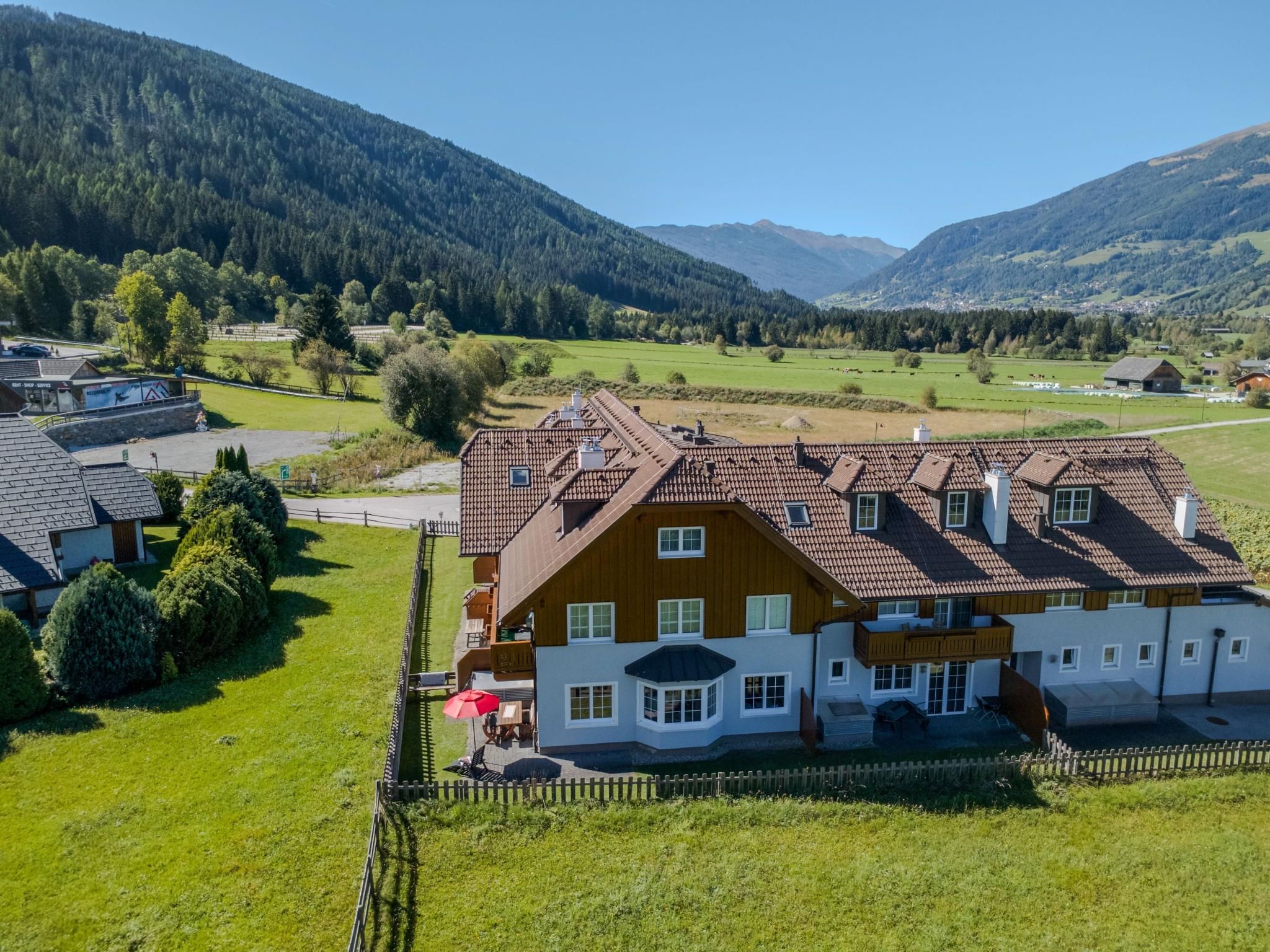 A large house with a sloped roof, surrounded by a fenced yard, set in a rural area with mountains in the background.