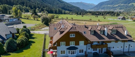 A large house with a sloped roof, surrounded by a fenced yard, set in a rural area with mountains in the background.