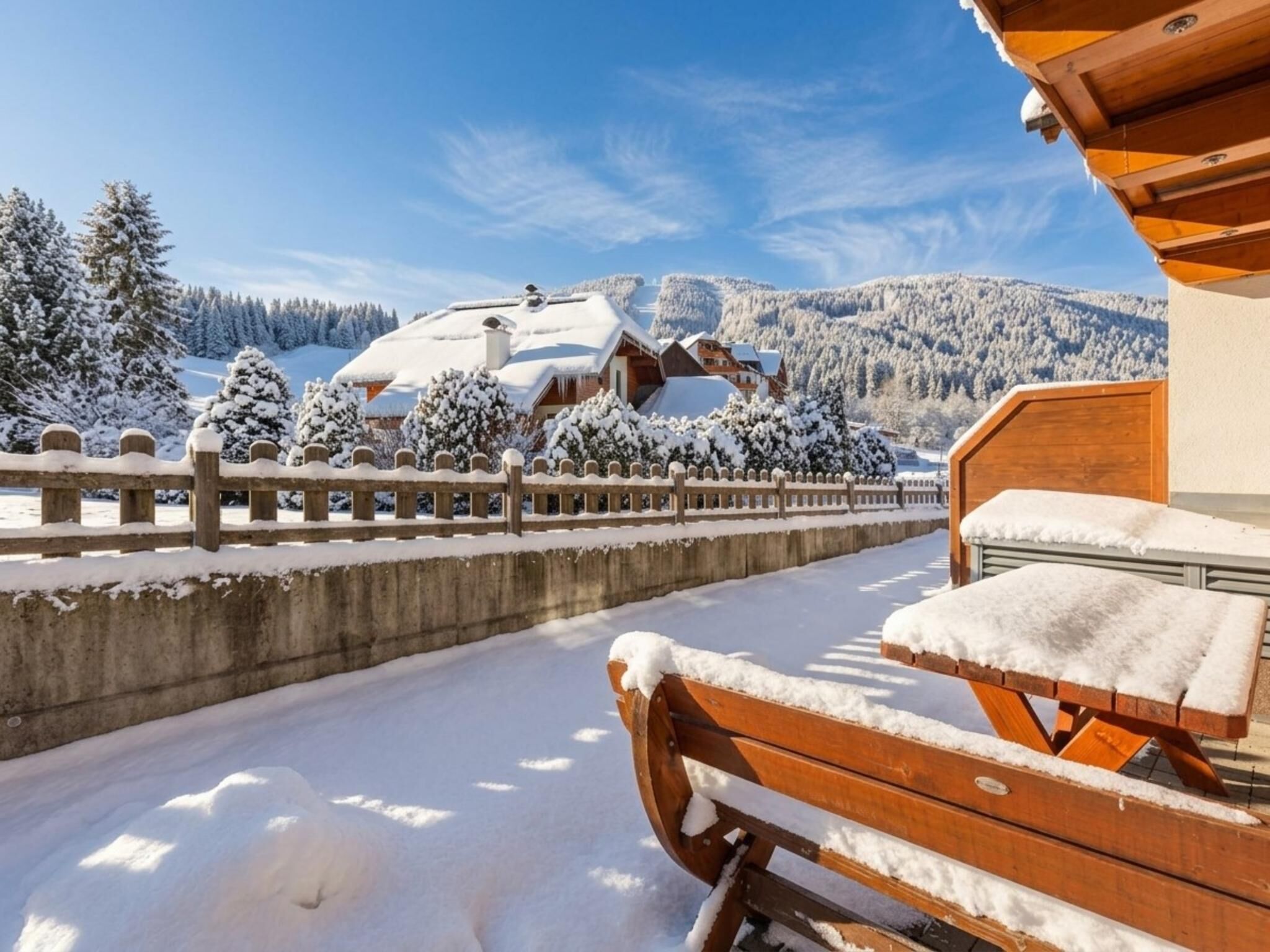 A wooden bench with snow on it, a concrete barrier, and a view of snow-covered houses and trees in the background.