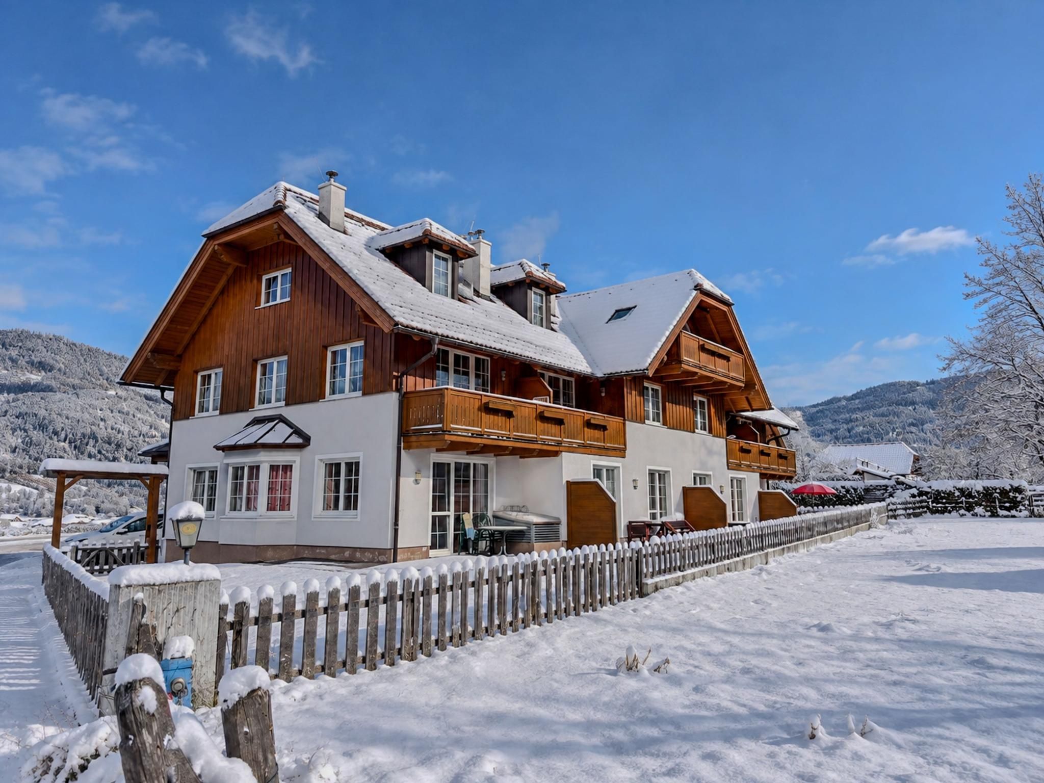 En tvåvånings chalet i trä med en balkong, en täckt uteplats och ett snötäckt landskap.