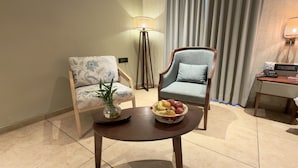 A hotel room with two armchairs, a small table with fruit, a lamp, and a desk with a phone.