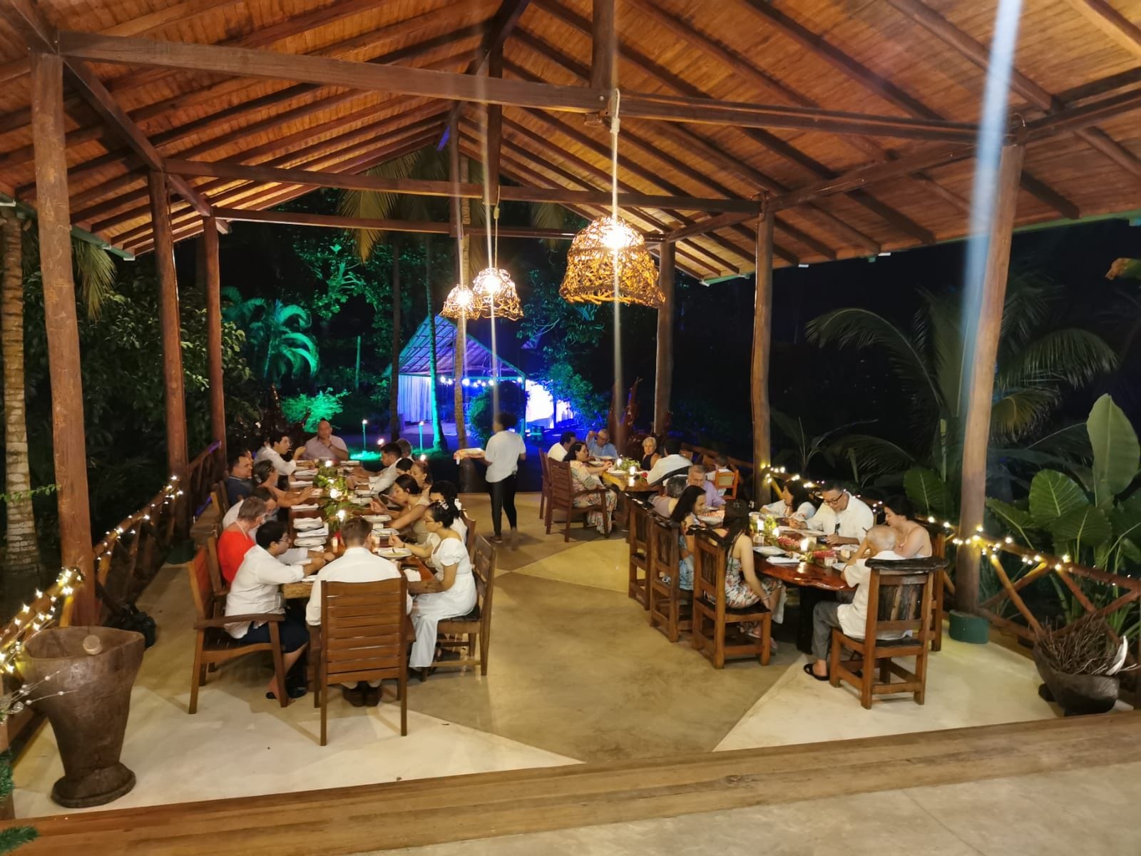 An outdoor dining area with wooden tables and chairs, surrounded by a thatched roof structure.