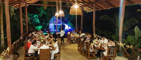 An outdoor dining area with wooden tables and chairs, surrounded by a thatched roof structure.