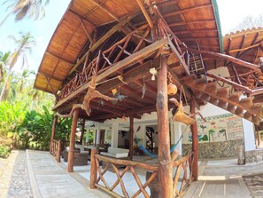 Una estructura de madera con una escalera que lleva a una zona cubierta, rodeada de follaje y un edificio con un mural.