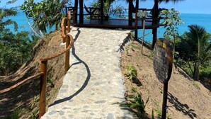 Una estructura con techo de paja, con un camino de piedra que la lleva hacia ella, rodeada de vegetación tropical y con vista a una masa de agua.