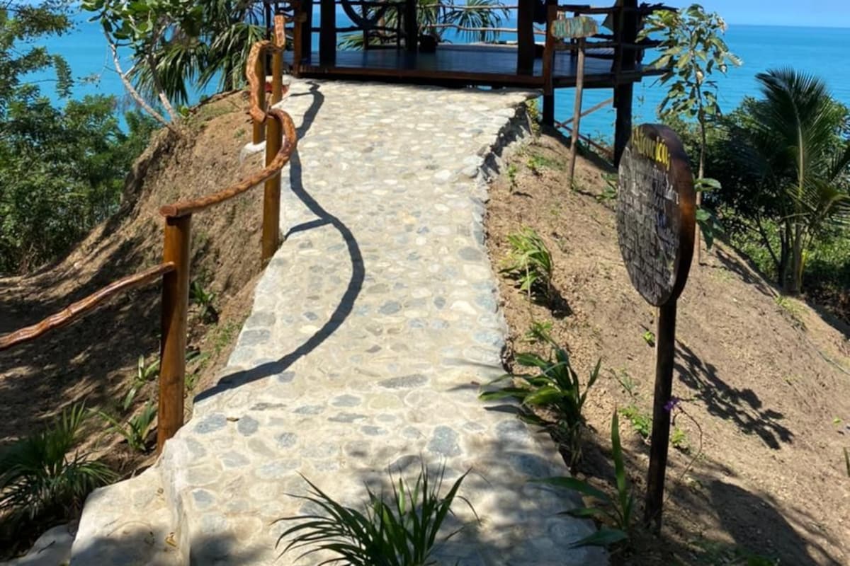 Una estructura con techo de paja, con un camino de piedra que la lleva hacia ella, rodeada de vegetación tropical y con vista a una masa de agua.