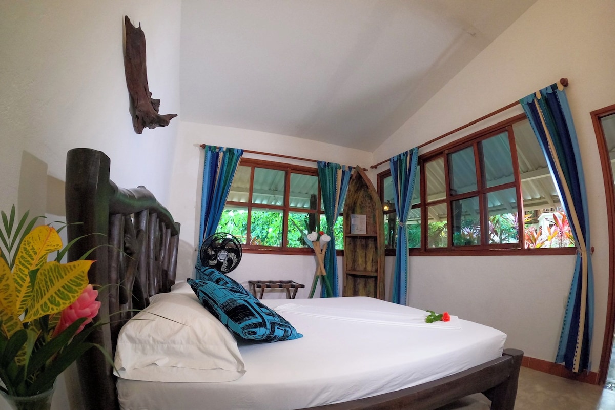 Una habitación con una cama, cabecera de madera, ventana con cortinas y un ventilador.