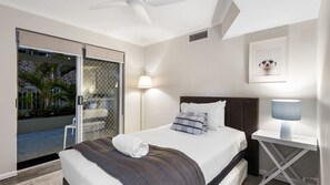 Ein Hotelzimmer mit Bett, Nachttisch, Lampe, Deckenventilator und einer Schiebeglastür, die auf einen Balkon mit Blick auf Palmen führt.