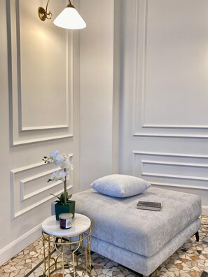 Une pièce avec un canapé gris recouvert de tissu, une petite table ronde avec un pied doré, un vase blanc orné de fleurs et une lampe fixée au mur.