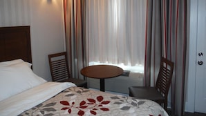 Ein Hotelzimmer mit einem Bett, einem runden Tisch, zwei Stühlen, einem Fenster mit Vorhängen und einer Wandleuchte.