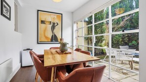 A dining area with a wooden table and brown chairs, a framed artwork above the table, and a view of a garden through large windows. - The Brick House East Melbourne (East Melbourne)