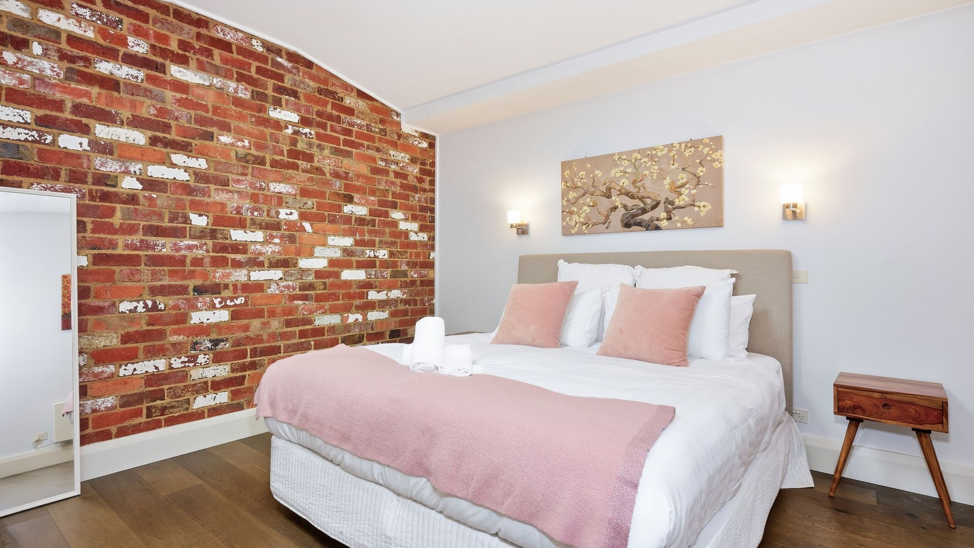 Une chambre avec un mur en briques, un lit avec une literie blanche et rose, une table de chevet et un luminaire fixé au mur.