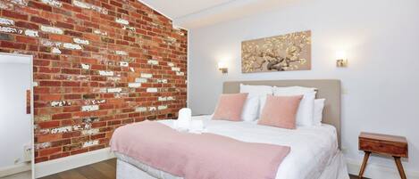 A bedroom with a brick wall, a bed with white and pink bedding, a nightstand, and a wall-mounted light fixture.