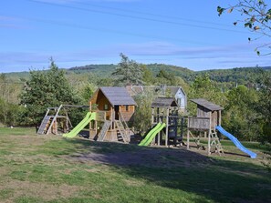 A playground with wooden play structures, including slides and climbing areas, set against a backdrop of green hills.