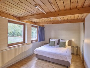 A bedroom with a wooden ceiling, a bed with white bedding, a bedside table with a lamp, and a window with a view of greenery.