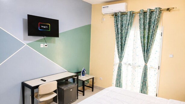 Et hotelværelse med en seng, et skrivebord med stol, et fjernsyn ophængt på væggen og en aircondition-enhed.