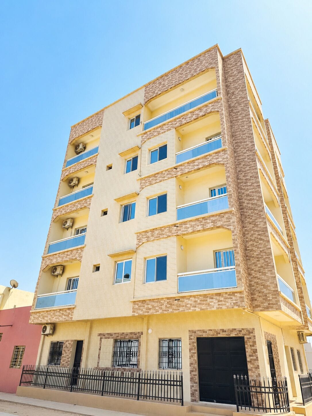 A multi-story building with balconies, air conditioning units, and a black gate at the entrance.