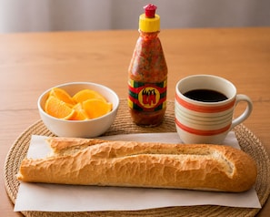 A baguette, a bowl of orange slices, a bottle of hot sauce, and a striped mug on a woven placemat.