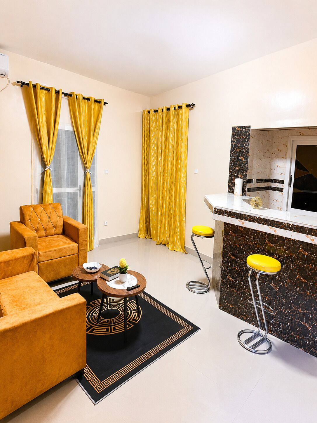A modern living room with a sofa, a coffee table, yellow curtains, a kitchen area with a bar stool, and a patterned rug.