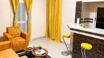 A modern living room with a sofa, a coffee table, yellow curtains, a kitchen area with a bar stool, and a patterned rug.