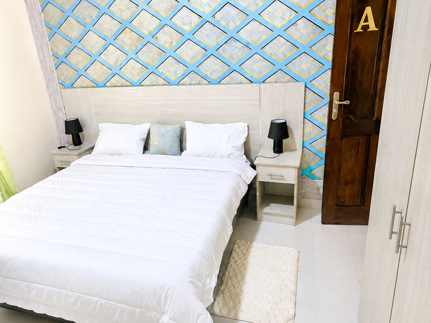 Un dormitorio con una cama, dos mesitas de noche, una mesita auxiliar, una puerta con la letra 'A' y un papel tapiz estampado.