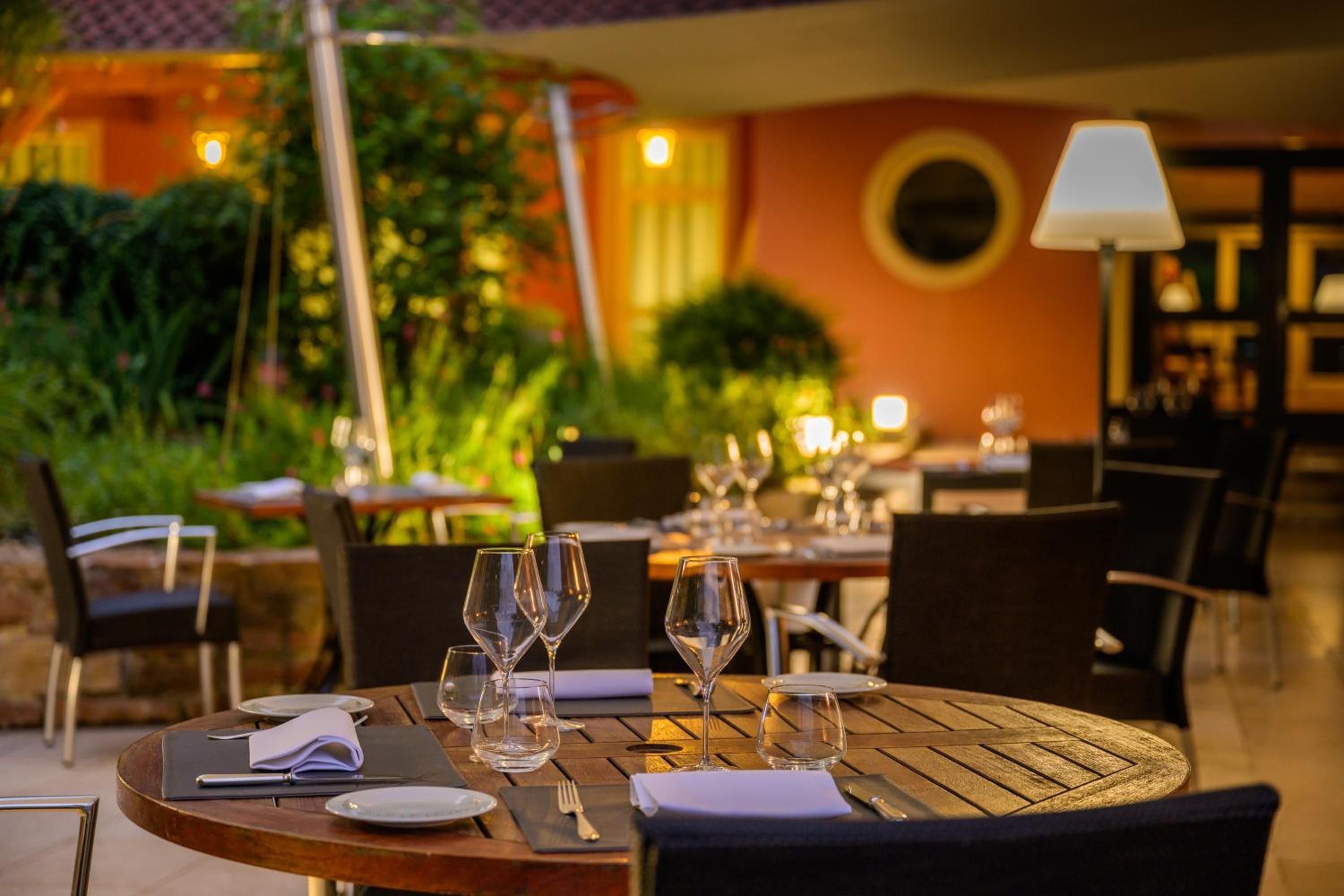 An outdoor dining area with tables set for two, featuring clear glassware and plates, surrounded by chairs and a lamp.