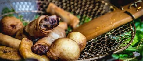 Uma cesta com diversos alimentos fritos, incluindo um item enrolado e vários itens redondos, sobre uma superfície com vegetação ao fundo.