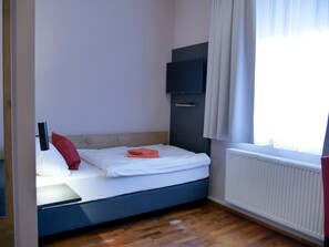 Una cama individual con colchón blanco y cabecera azul, un televisor de pantalla plana montado en la pared y una ventana con cortinas.