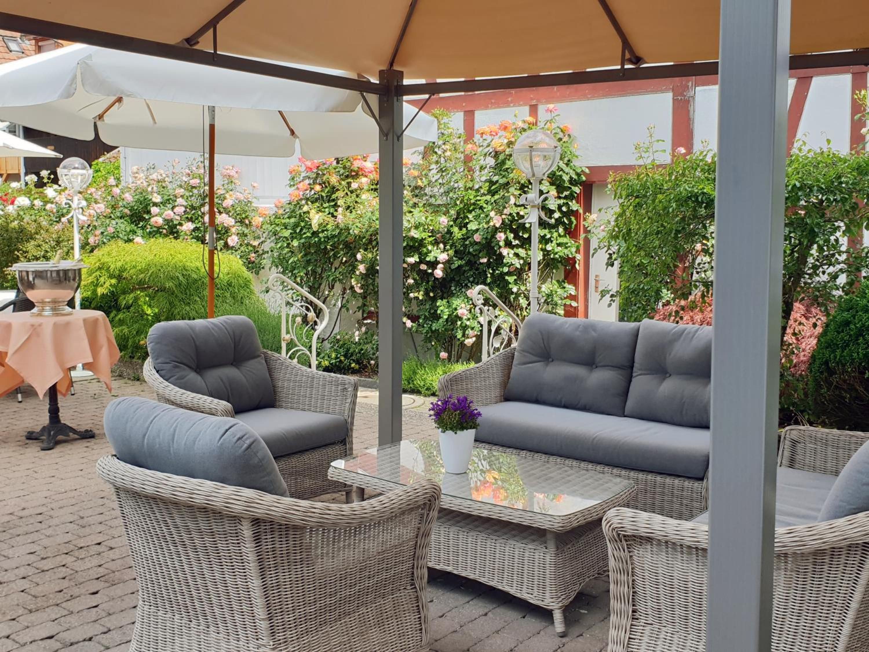 An outdoor seating area with wicker furniture and a table, surrounded by greenery and flowers.