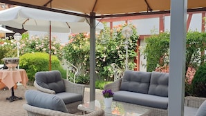An outdoor seating area with wicker furniture and a table, surrounded by greenery and flowers.