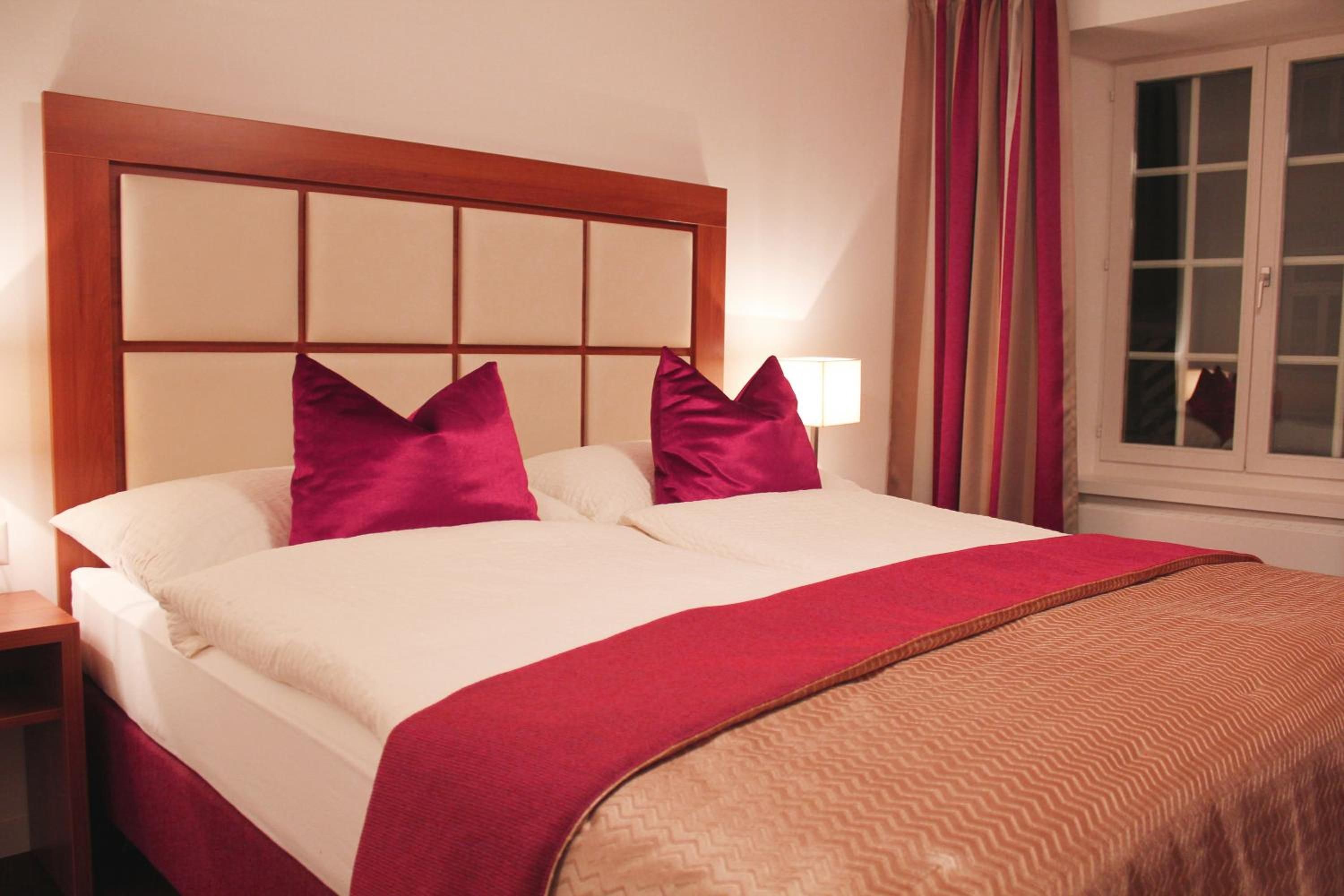 A hotel room with a large bed, a wooden headboard, two pillows, a bedside lamp, and a window with curtains.