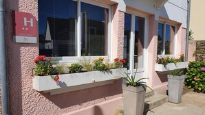 Un edificio con fachada rosa, un letrero con la letra 'H' que indica un hotel, y macetas con diversas plantas y flores.