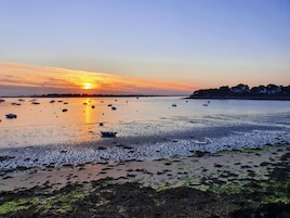 A coastal scene with boats on calm water, a sunset, and a shoreline with vegetation.