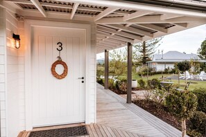 Una puerta blanca con el número 3 y un adorno, un porche cubierto con vista a una casa y jardín, y una terraza de madera.
