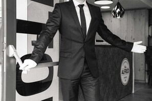 Una persona in abito che si trova in un corridoio con una reception con la scritta 'ALFRED' e un design d'interni moderno.