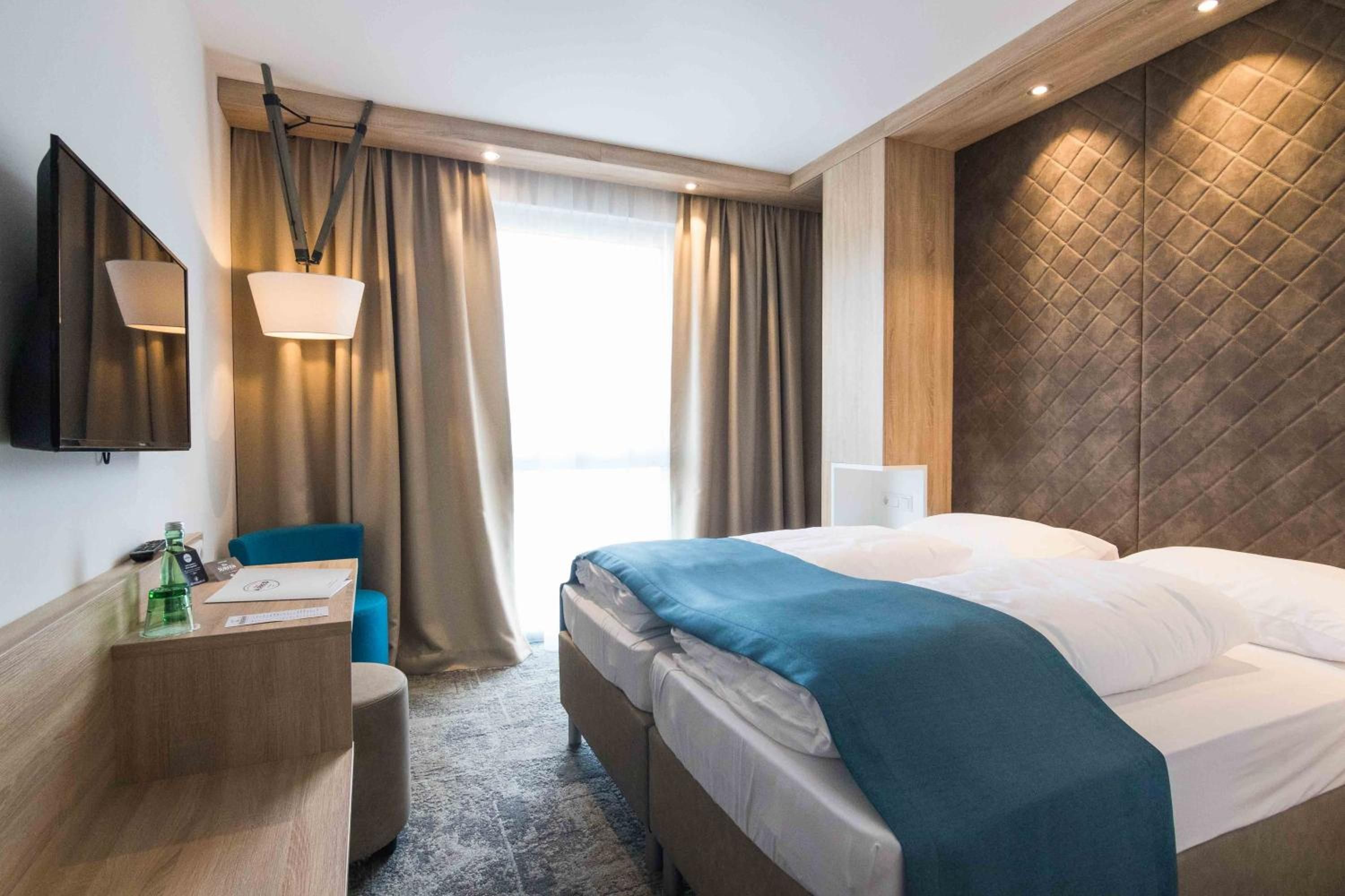 Habitación de hotel con cama, escritorio, silla, televisor y ventana con cortinas.