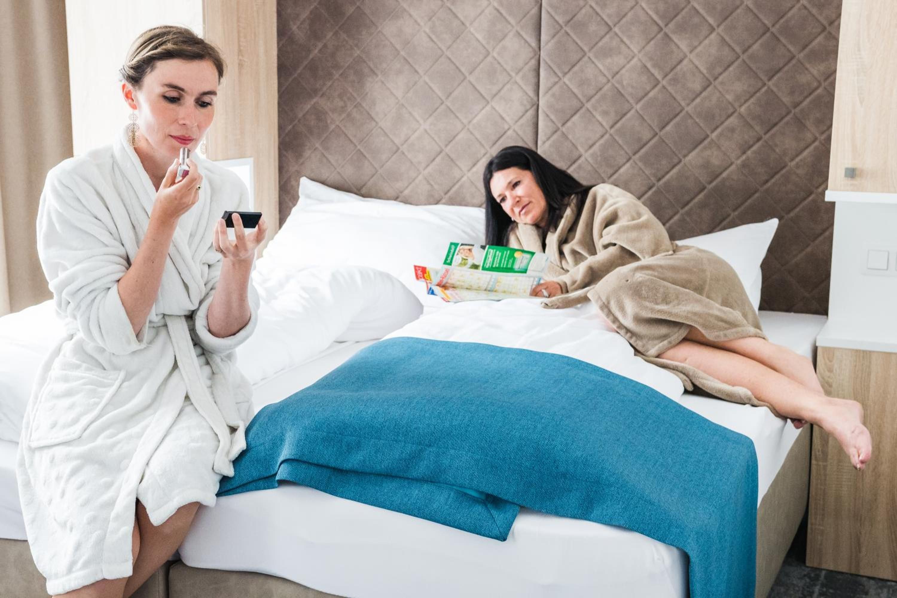 Una habitación de hotel con una cama, almohadas, una manta azul y una persona leyendo una revista.