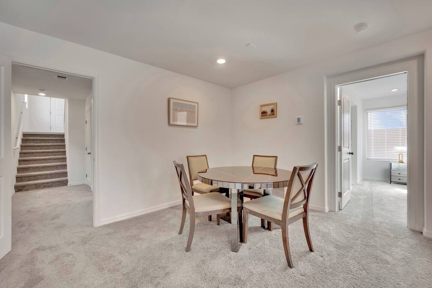 A room with a round table and four chairs, carpeted floor, white walls, and a staircase in the background.