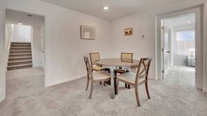 Quarto com mesa redonda e quatro cadeiras, piso coberto por carpete, paredes brancas e escada ao fundo.
