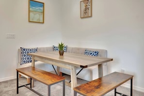 Un coin repas avec une table et des bancs en bois, un canapé agrémenté de coussins à motifs et deux œuvres d’art encadrées au mur.