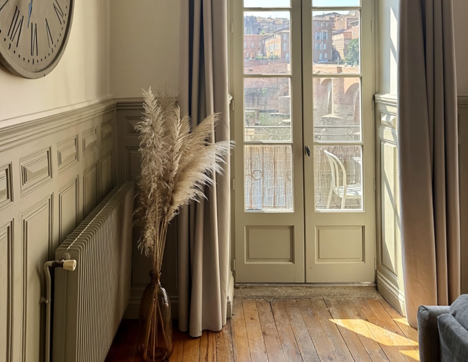 Une pièce avec un radiateur, un vase contenant des plumes d’herbe des pampas, une porte donnant sur des immeubles et une chaise.
