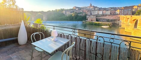 A terrace with a table and chairs overlooking a river, with buildings in the background.