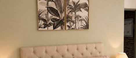 A room with a tufted headboard, two pillows, a wall-mounted plant artwork, and a lamp on a side table.