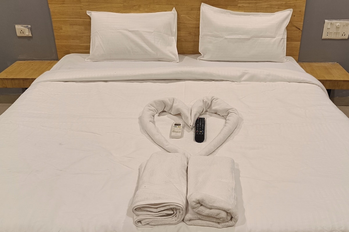 Een netjes opgemaakt bed met witte lakens, een opgevouwen handdoek aan het voeteneind en een afstandsbediening op een handdoek in het midden.